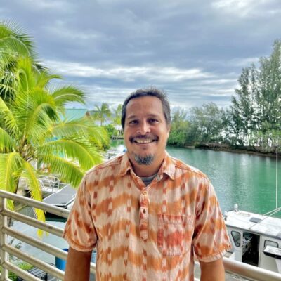 A smiling man in a patterned shirt stands by water, boats, and palm trees under a cloudy sky.
