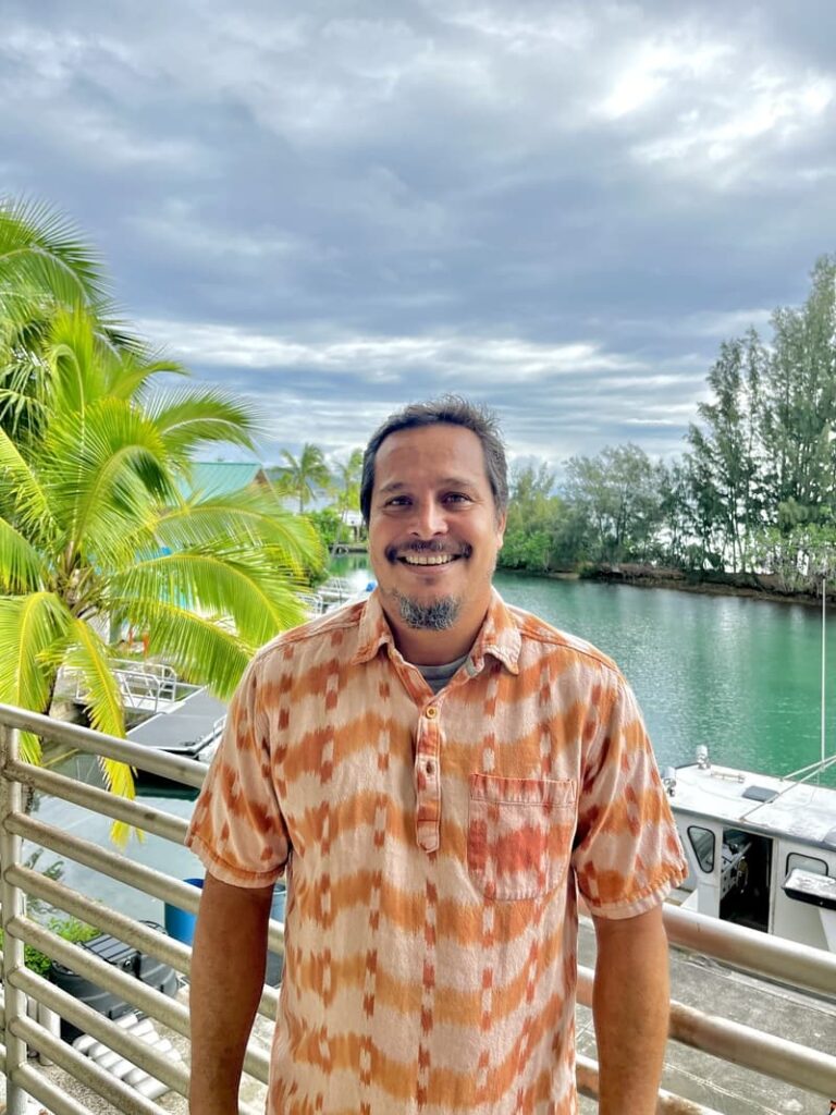 A smiling man in a patterned shirt stands by water, boats, and palm trees under a cloudy sky.