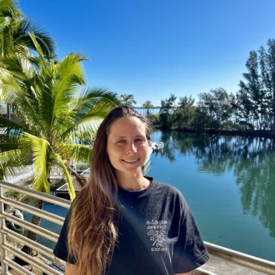 Smiling woman with long hair stands by a railing near water, palm trees, and a clear blue sky.