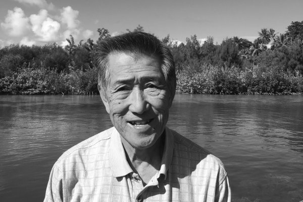 Older man smiling by a river, with trees and cloudy sky in the background; black-and-white photo.