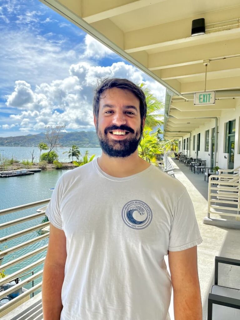 Smiling man with dark hair and beard stands on a balcony by water with mountains and palm trees in the background.