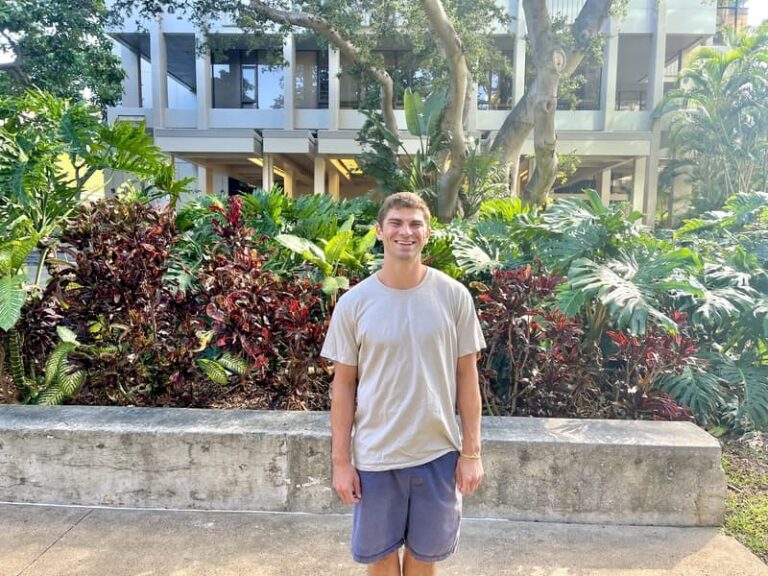 A young man in a beige shirt and shorts stands smiling in front of lush greenery and a modern building.