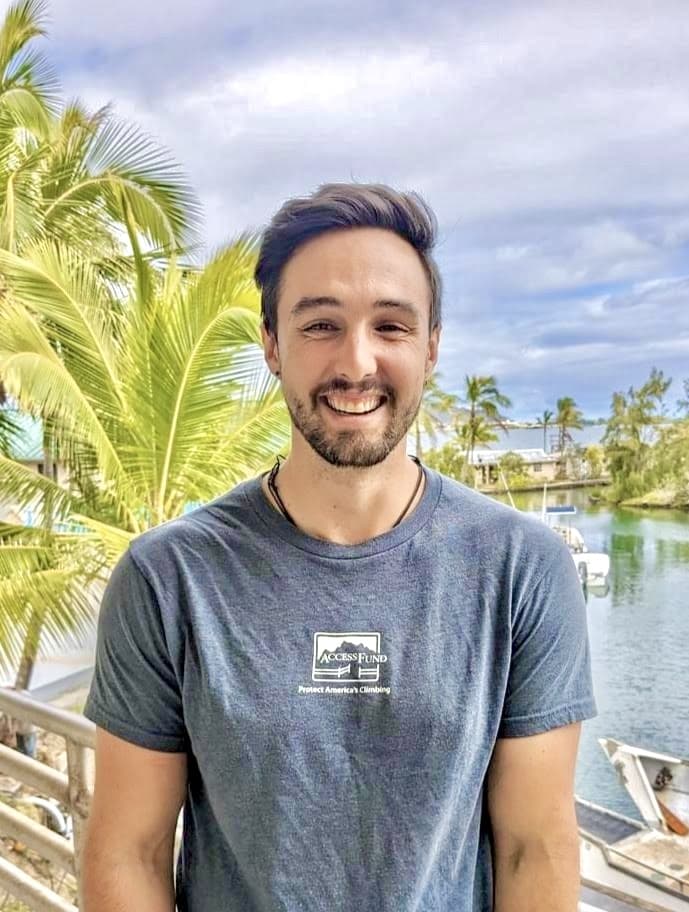 A smiling man stands outdoors by water and palm trees, wearing a grey T-shirt with a printed logo.