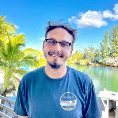 Man with glasses smiling outside by water, palm trees, and a blue sky with clouds, wearing a blue t-shirt.
