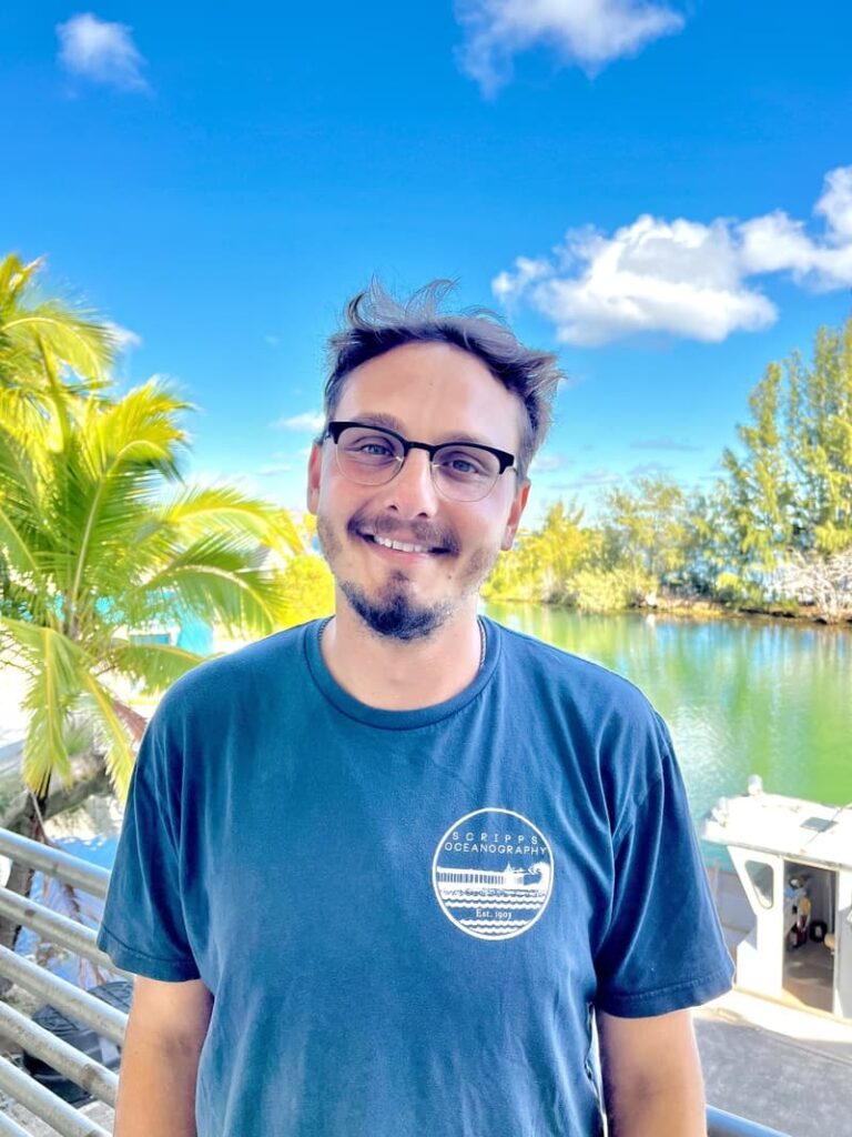Man with glasses smiling outside by water, palm trees, and a blue sky with clouds, wearing a blue t-shirt.