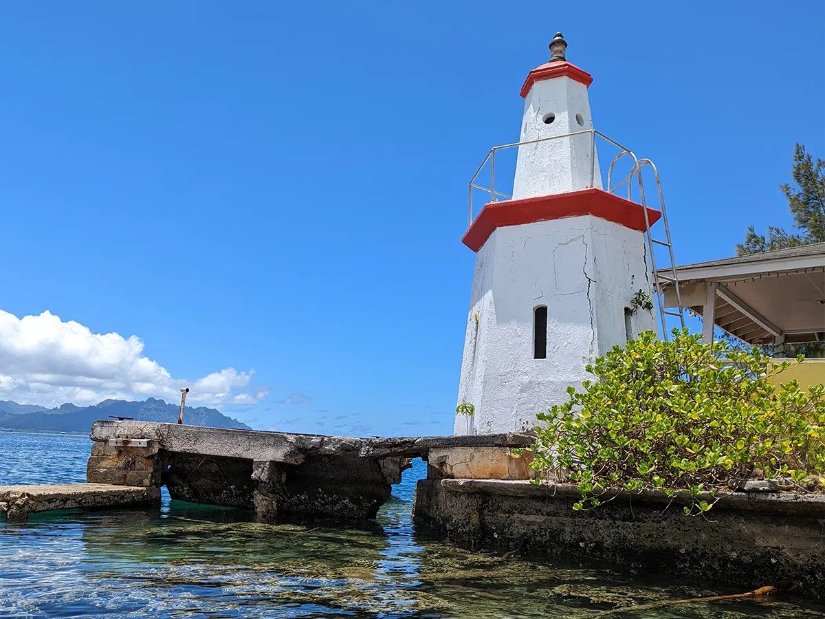 A white lighthouse with red trim stands by clear water under a bright blue sky, with greenery and mountains nearby.