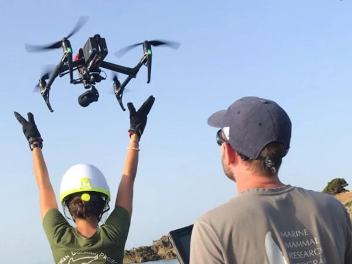 Two researchers operate a drone; one raises arms while the other monitors with a laptop outdoors.