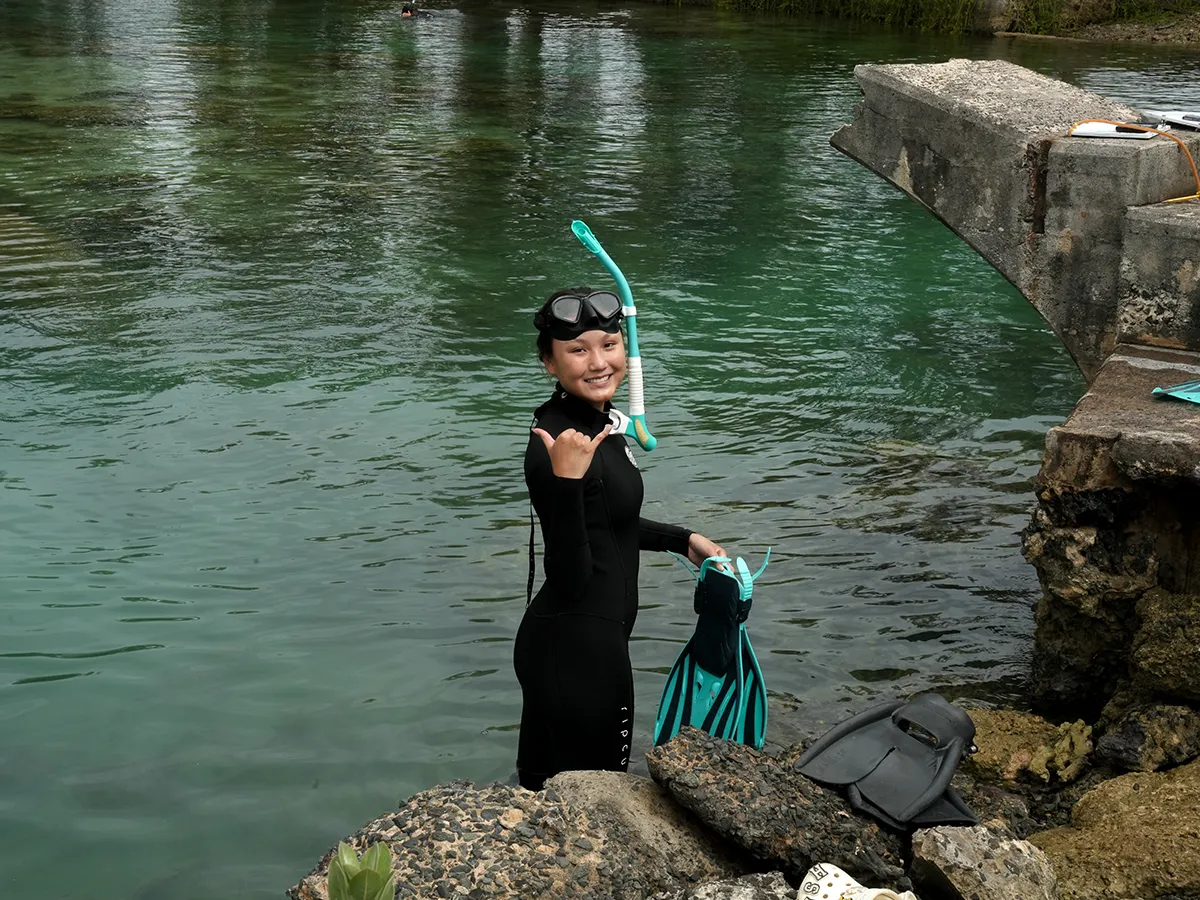 A person in a wetsuit stands by the water, smiling and giving a shaka sign, holding snorkeling gear.