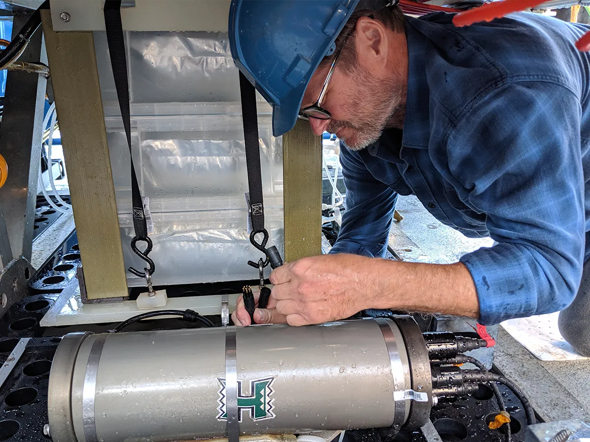 A man in a blue hard hat works on marine research equipment under a structure.