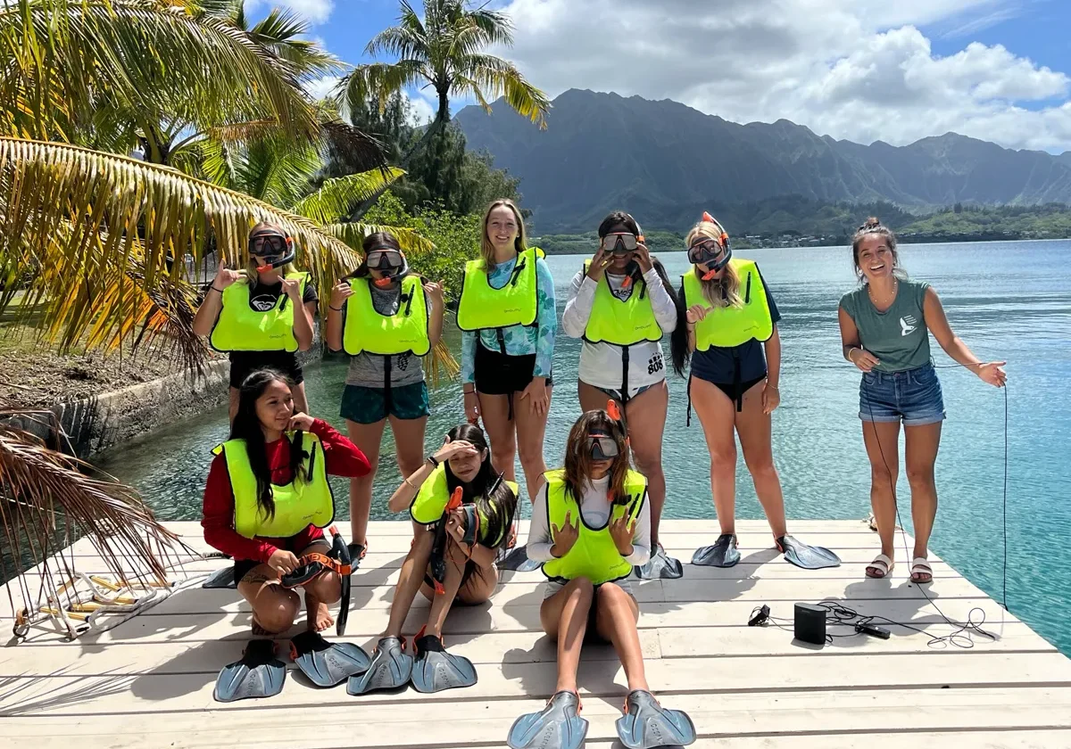 A group of people in snorkel gear poses on a dock by the water with mountains and palm trees in the background.