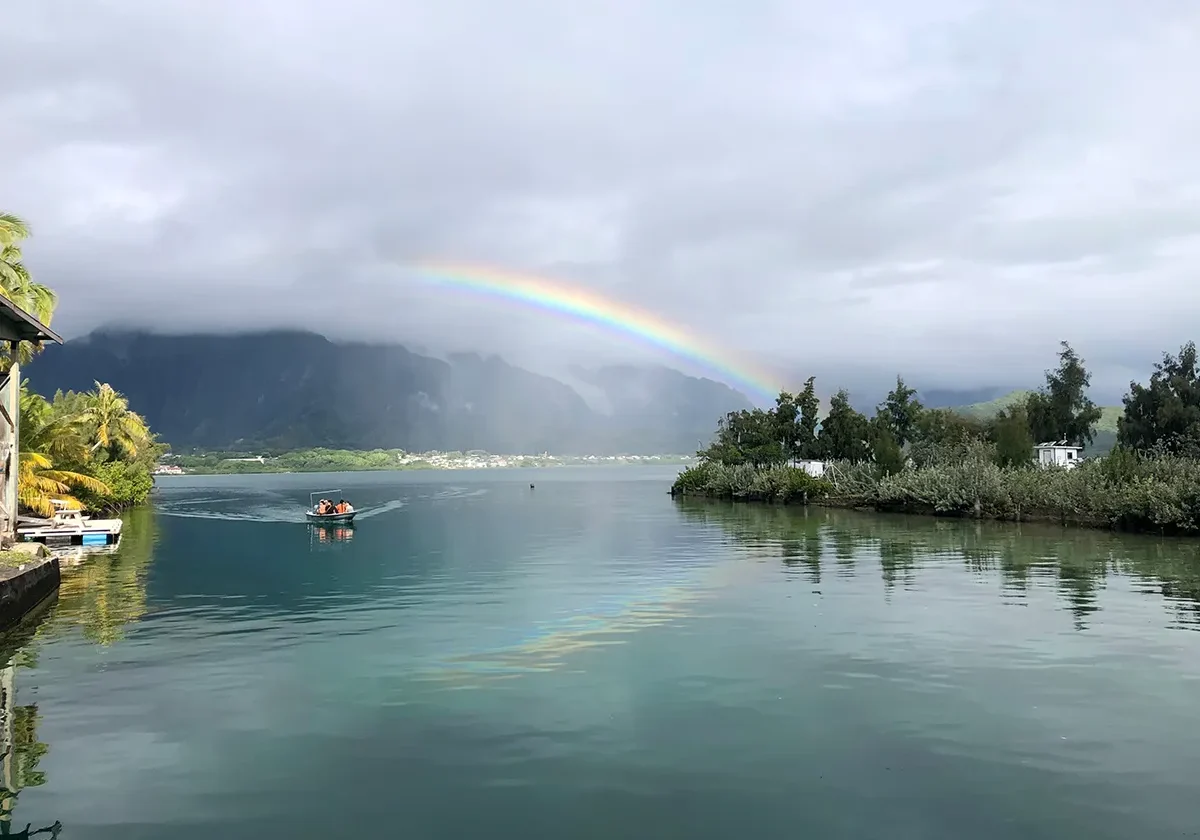 A small boat on a calm lake with a rainbow arching over mountains under a cloudy sky.