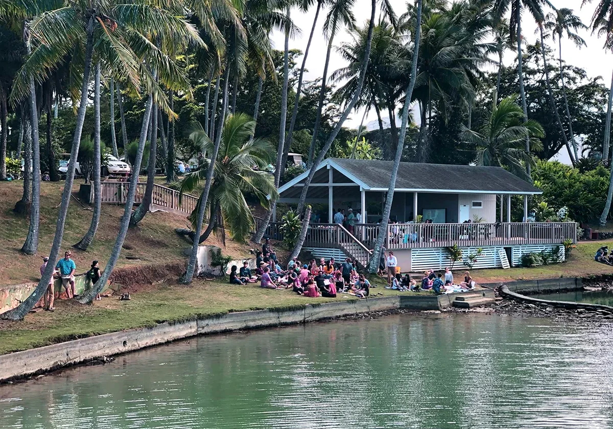 People sitting and standing by a lakeside building surrounded by palm trees on a sunny day.