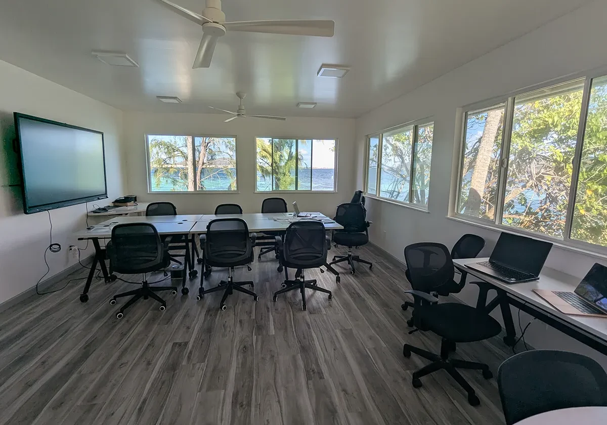A modern conference room with chairs, tables, laptops, large windows, and a view of trees and water outside.