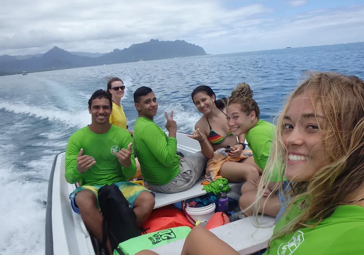 Six smiling people in green shirts ride a small boat on the ocean with mountains in the background.