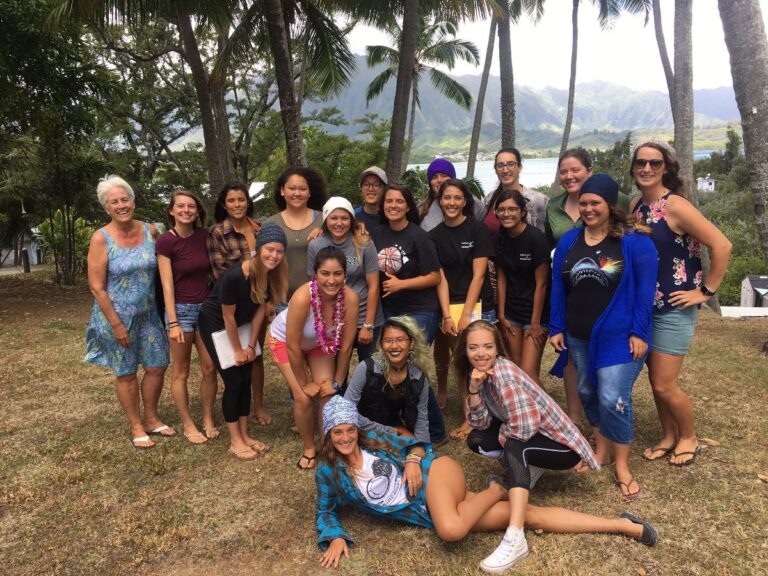 A diverse group of people pose and smile outdoors in front of palm trees and mountains.