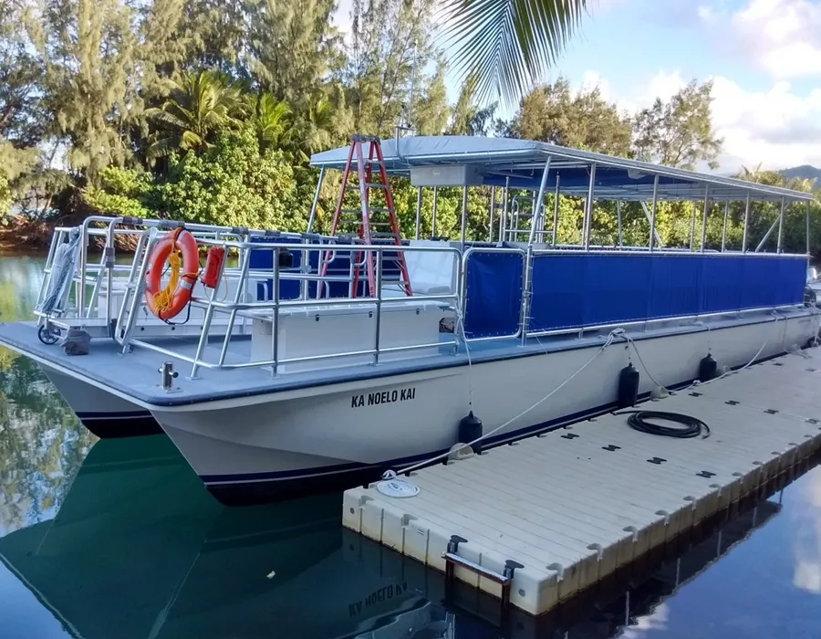 A white catamaran named "Ka Noelo Kai" docked by a floating pier, surrounded by trees and water.