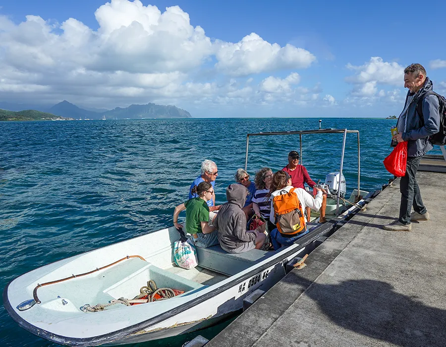 A group of people sit in a small boat at a dock, with one person standing nearby, ocean and mountains in background.
