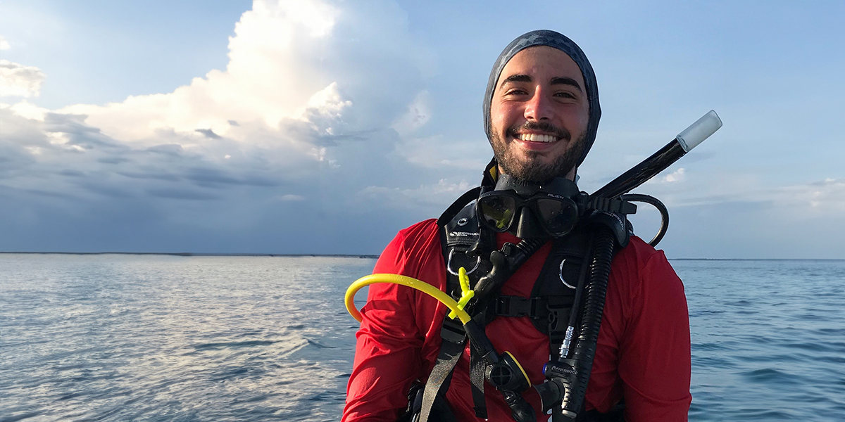 A smiling scuba diver in red gear sits on a boat with the ocean and cloudy sky in the background.
