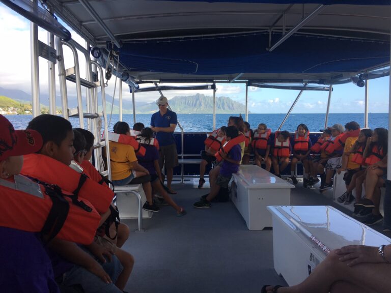 Children wearing life jackets sit on a boat, listening to an adult guide, with mountains visible in the background.
