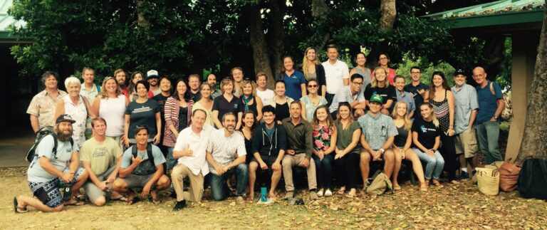 A large group of people poses together outdoors under trees, smiling for a group photo.