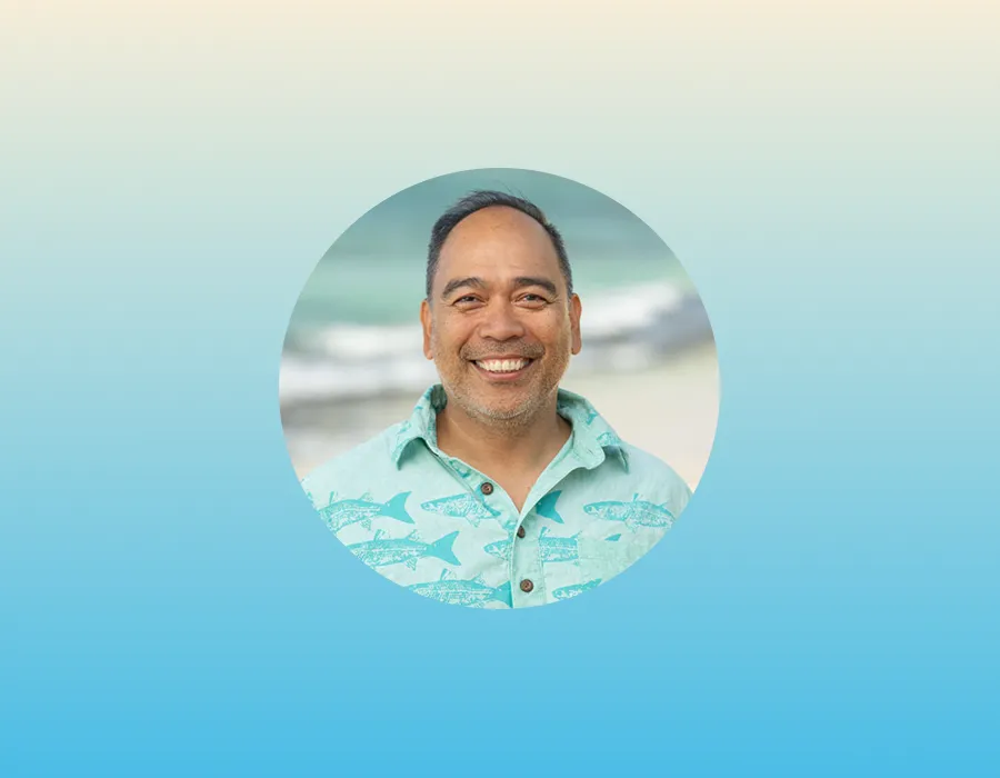 Smiling man in a fish-print shirt with a beach background, centered on a blue gradient background.