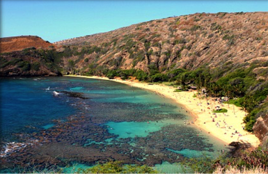 Curved sandy beach with turquoise water and rocky hills, bordered by lush greenery and scattered visitors.
