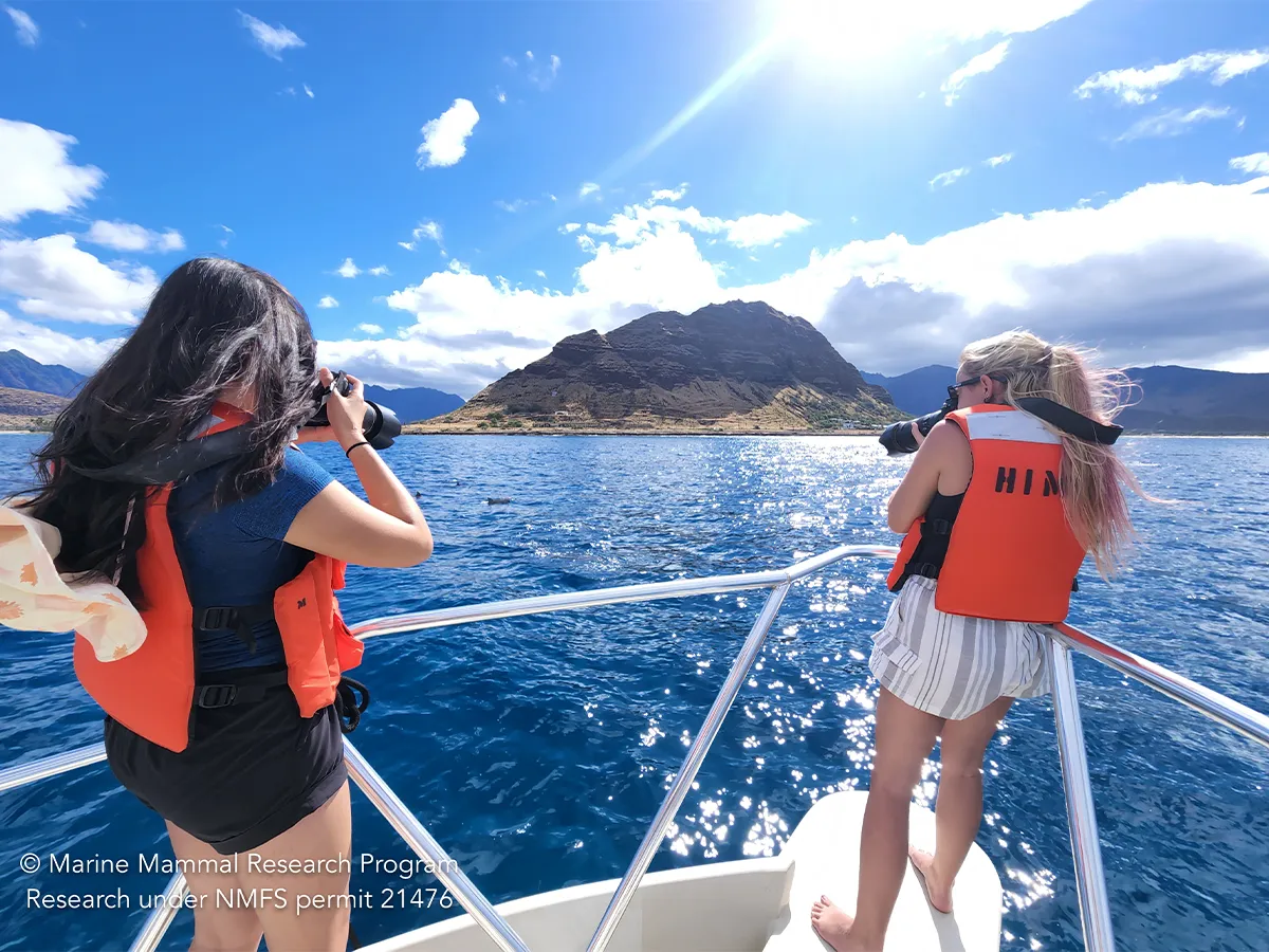 Two women in life vests photograph a mountainous island from a boat on clear blue water under a sunny sky.