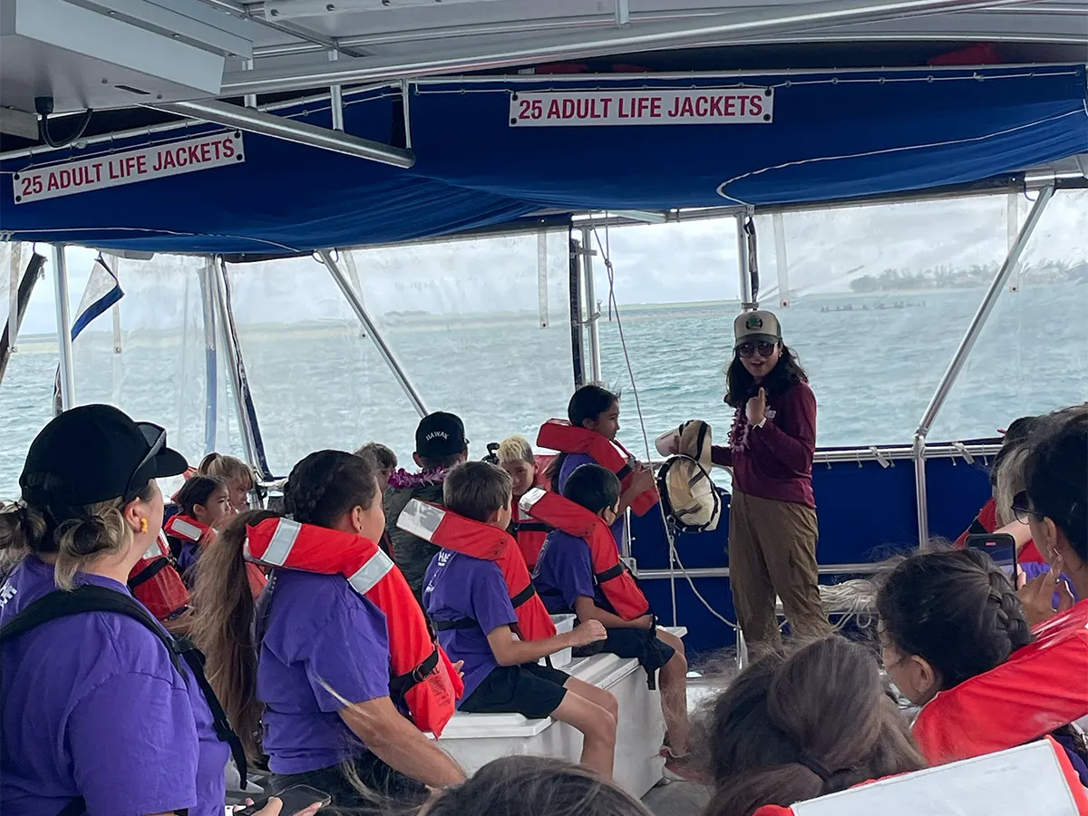 A group of children in life jackets listen to a woman speaking on a boat, with water visible outside.