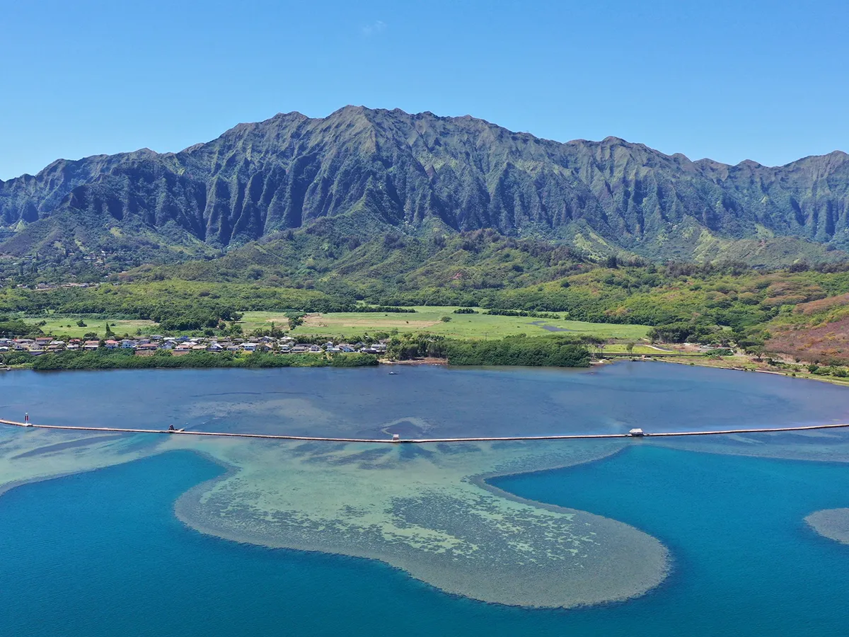 A long bridge crosses blue water with green mountains and a small town in the background under a clear sky.