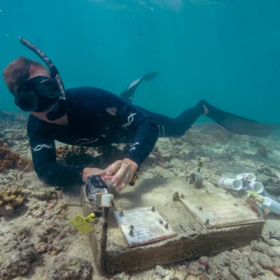 A scuba diver works with equipment on the ocean floor near some coral, with another diver in the background.
