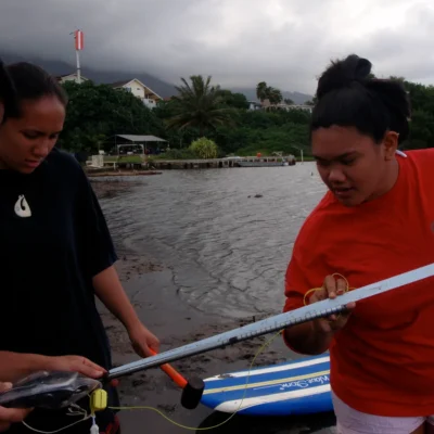 Three people by the water examine a long measuring tool, with trees and houses in the background.