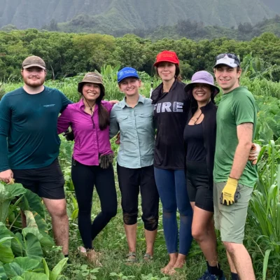 Six people stand together smiling in a lush, green field with mountains and trees in the background.