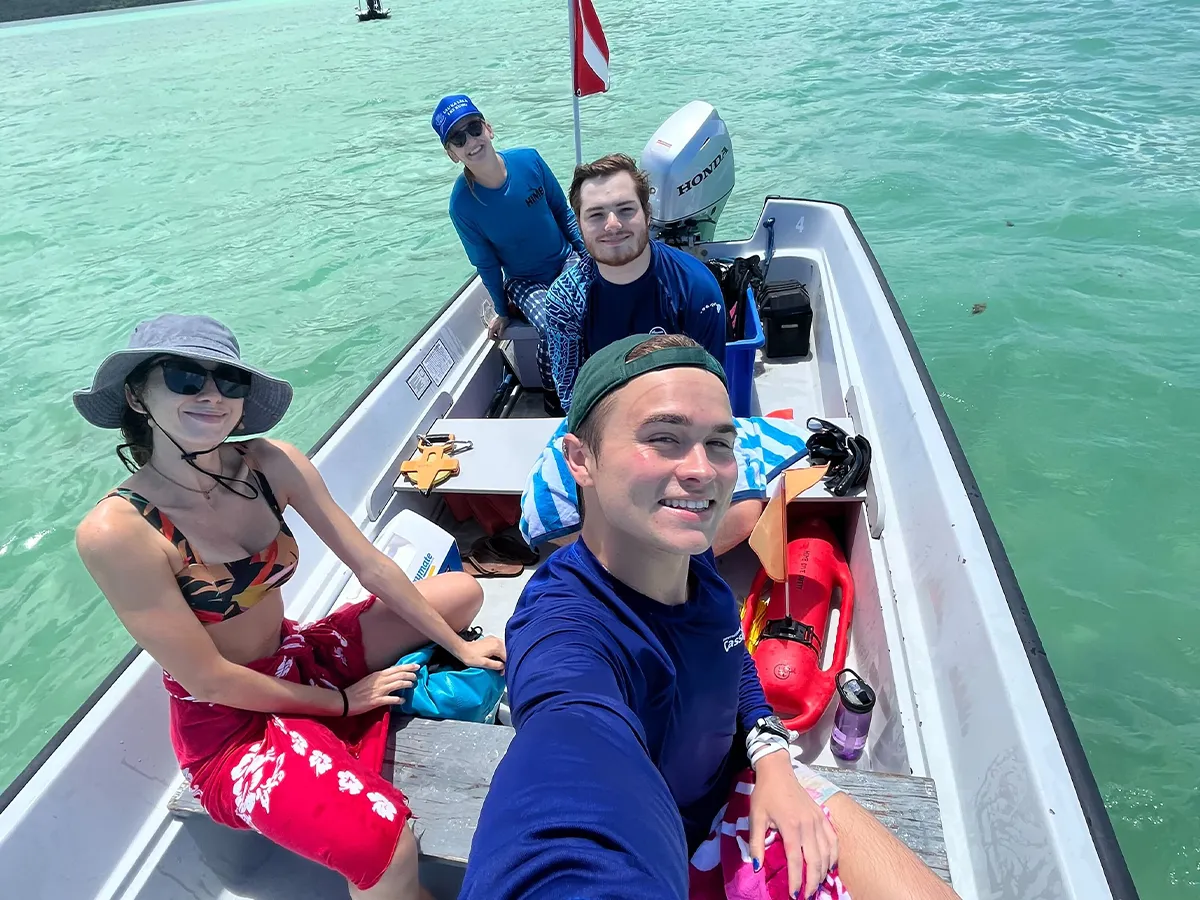 Four people smiling on a small boat in clear turquoise water, wearing hats and swimwear on a sunny day.