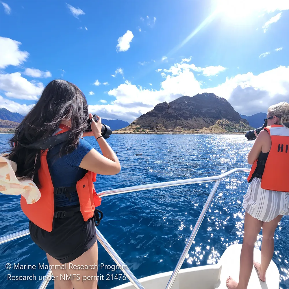 Two women in orange life vests photograph a scenic island from a boat on bright blue water under a sunny sky.