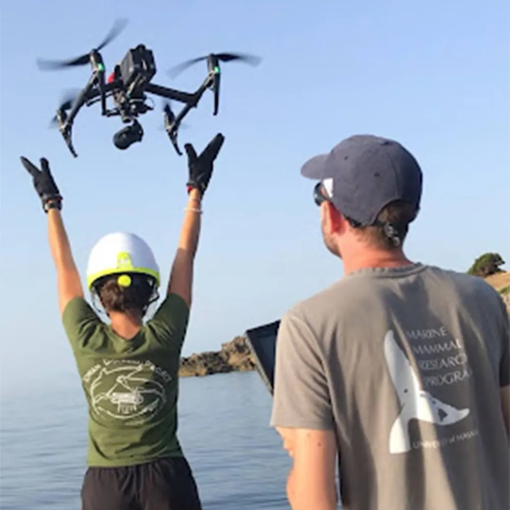 Two people operate a drone by the water; one raises arms as the drone hovers overhead on a clear day.