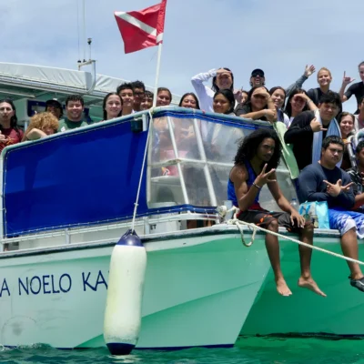 A group of people smile and pose on a boat named "KA NOELO KA" in turquoise water under a clear sky.