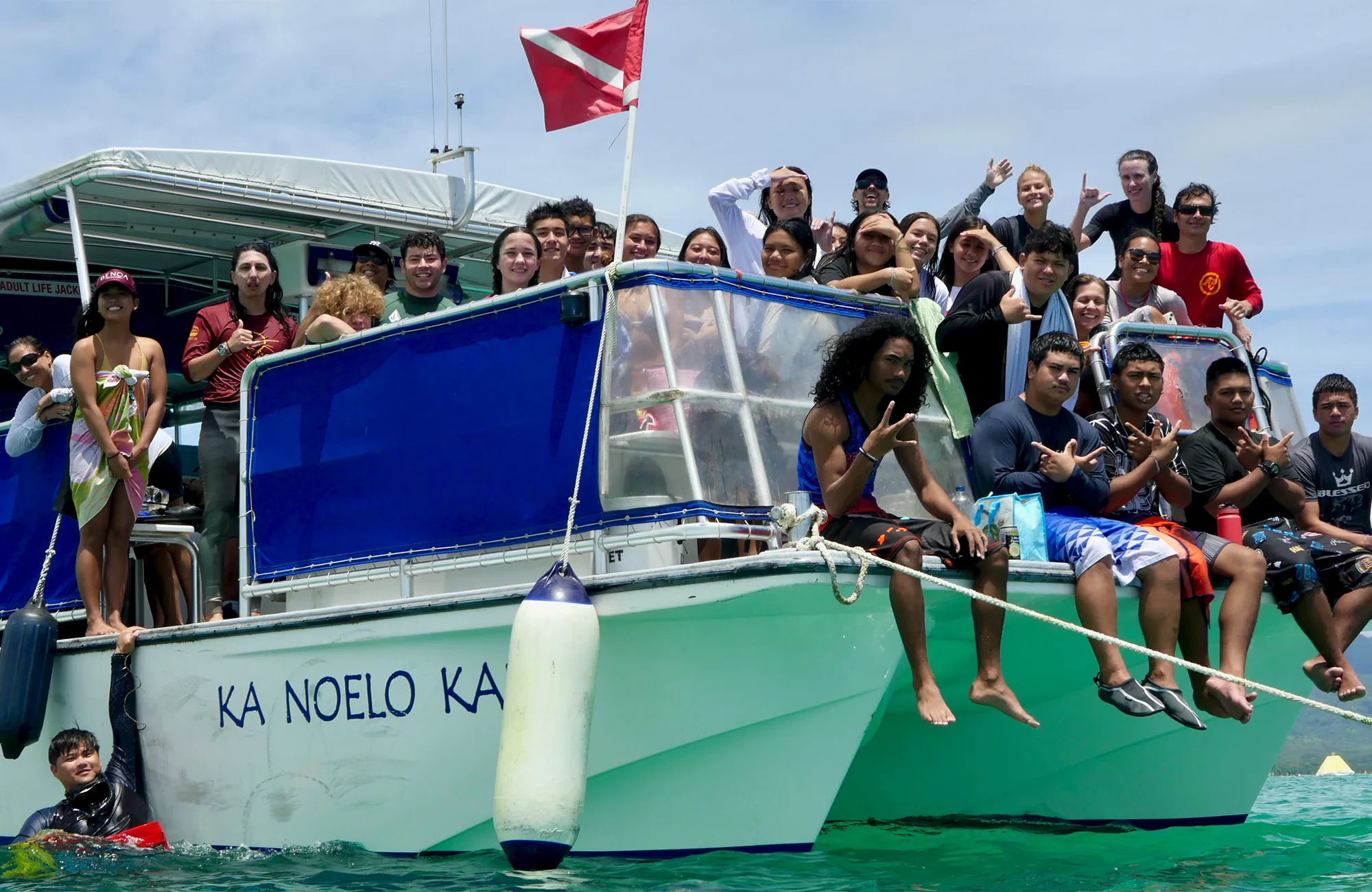 A group of people smile and pose on a boat named "KA NOELO KA" in turquoise water under a clear sky.
