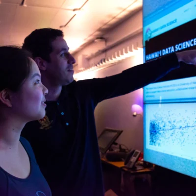 Two people look at and discuss data visualizations on a large screen in a tech-focused room.