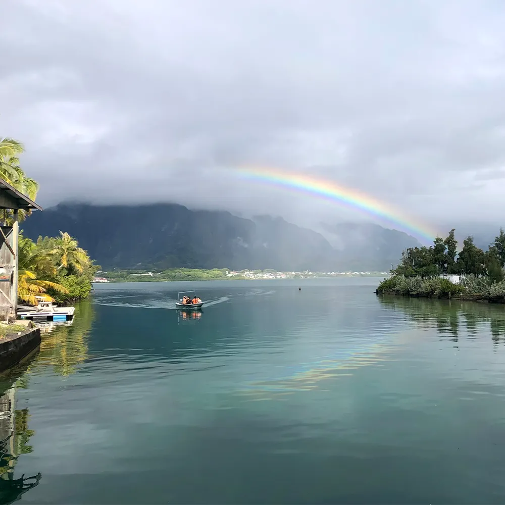 Small boat on calm water, tropical trees, mountains, and a rainbow under a cloudy sky in the distance.