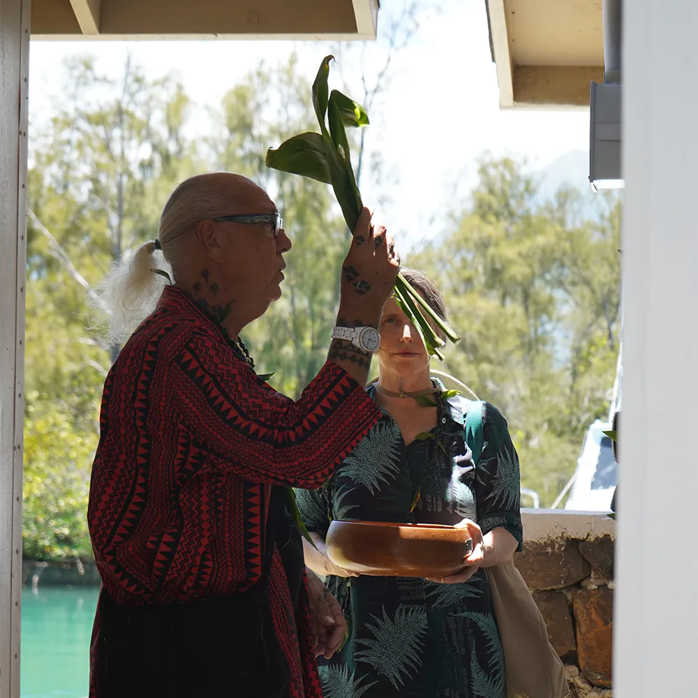 A man holds leafy plants while a woman holds a wooden bowl outdoors, both dressed in patterned clothing.