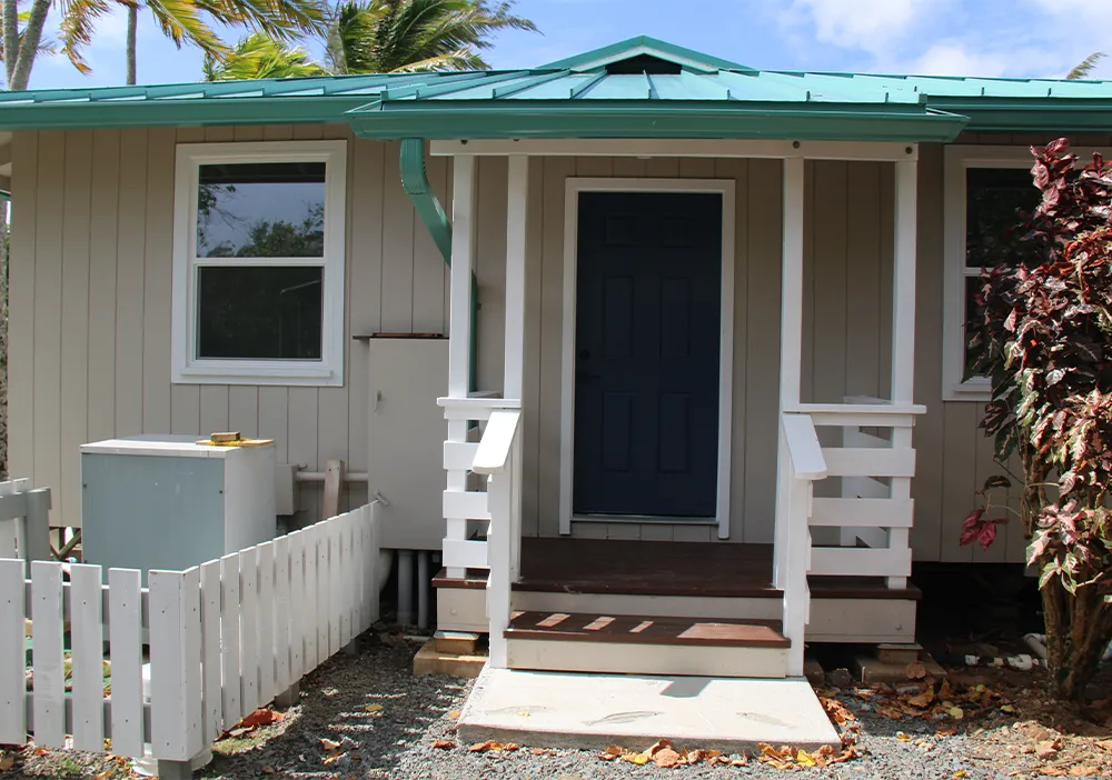 Small house with a teal roof, beige siding, and a dark blue front door, beside a white picket fence.