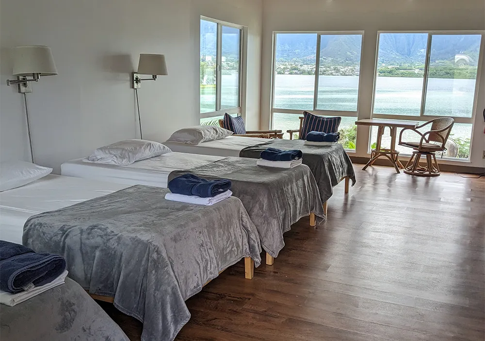 A bright room with three single beds, gray blankets, and large windows overlooking a lake and mountains.