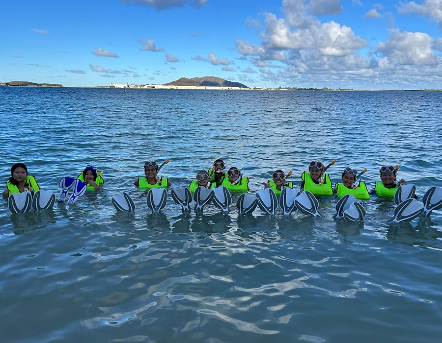 A group of kids in snorkel gear and life vests float in the ocean, holding flippers above the water.