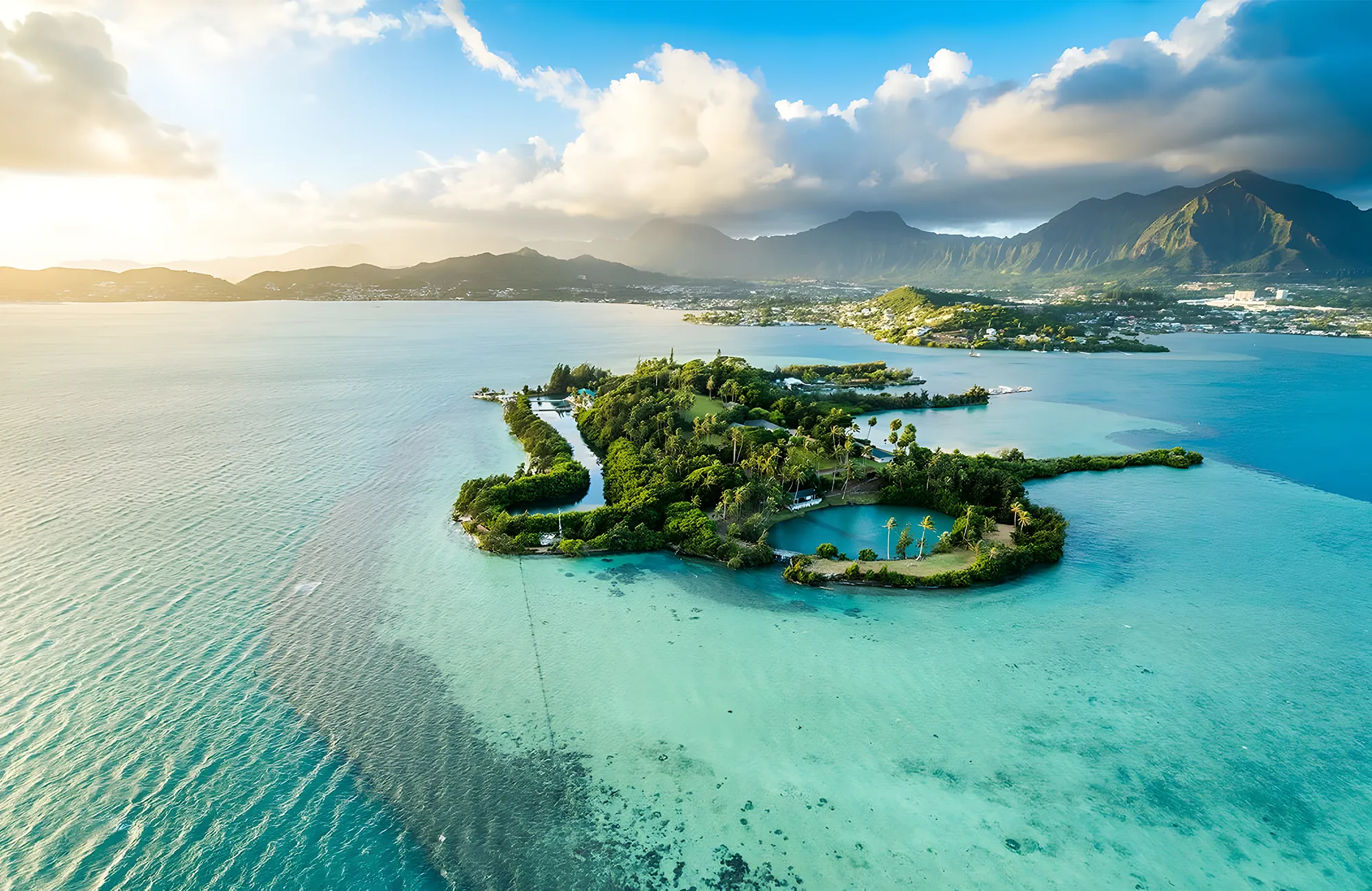 Aerial view of a lush green island surrounded by clear turquoise water and distant mountains under a sunny sky.