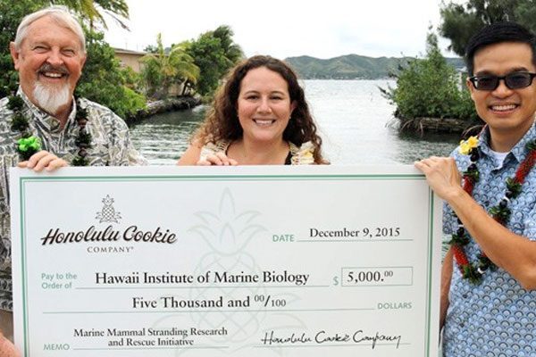 Three people hold a large $5,000 check for the Hawaii Institute of Marine Biology by a waterfront.