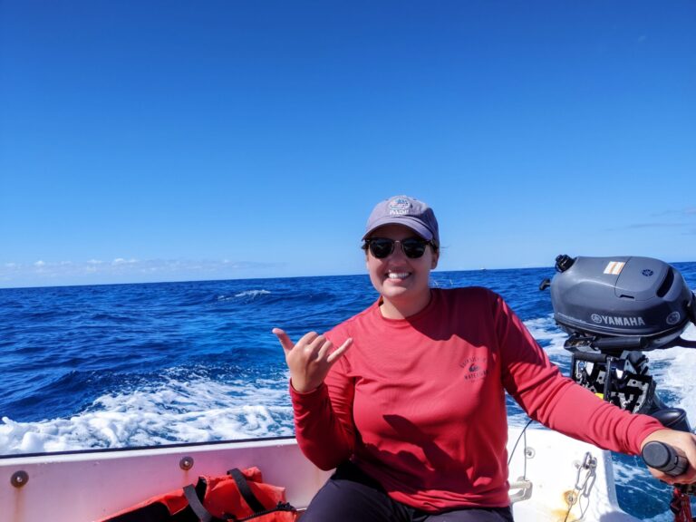 Smiling person in a red shirt and hat makes a shaka sign while riding a boat on the ocean under a blue sky.