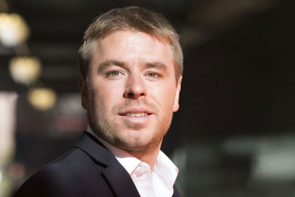 A man in a suit smiles confidently while standing outdoors with blurred lights in the background.