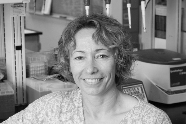 A woman with curly hair smiles in a laboratory setting with equipment in the background.