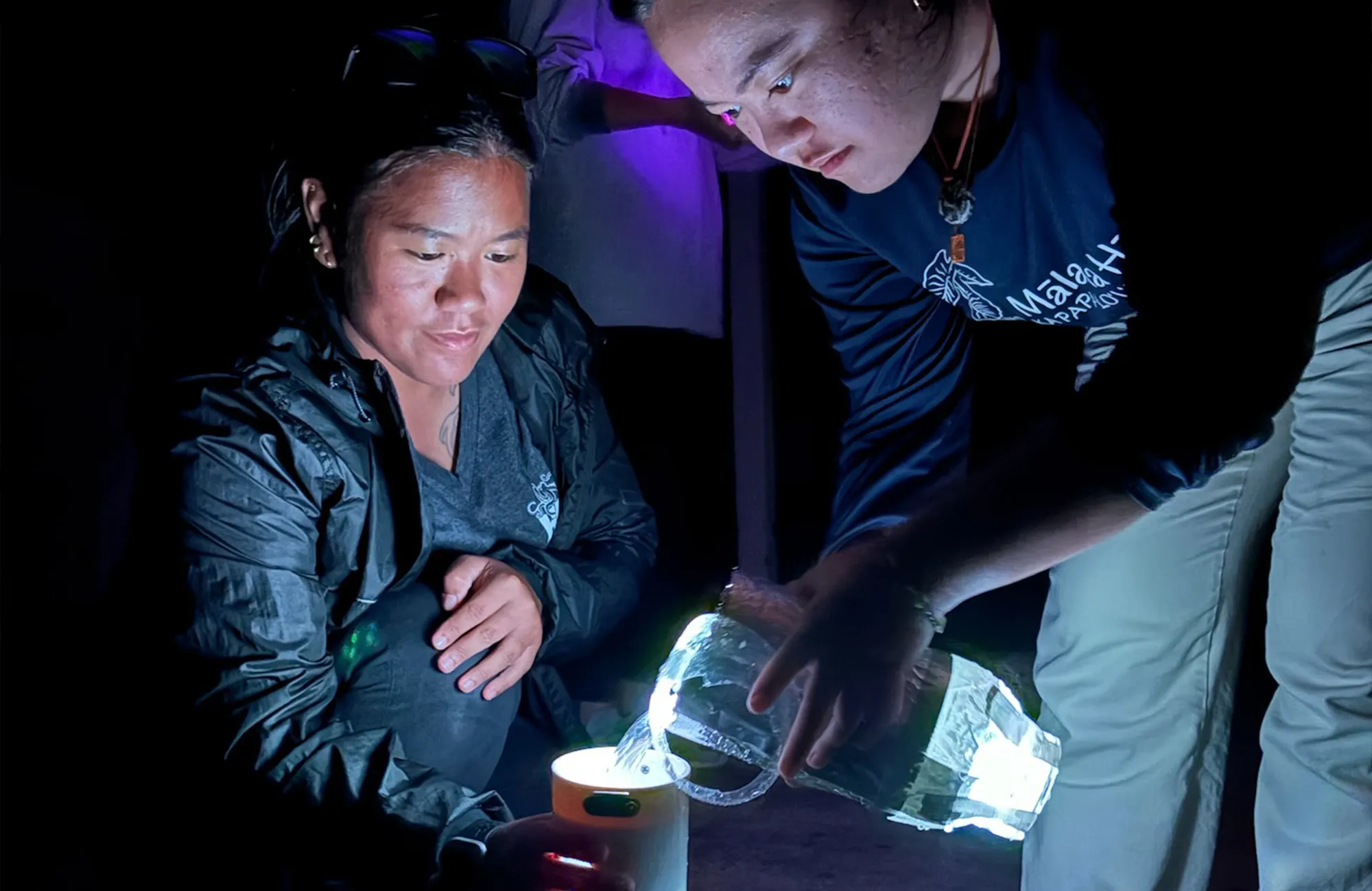 Two women pour water into a device, illuminated by a bright light in a dark setting.