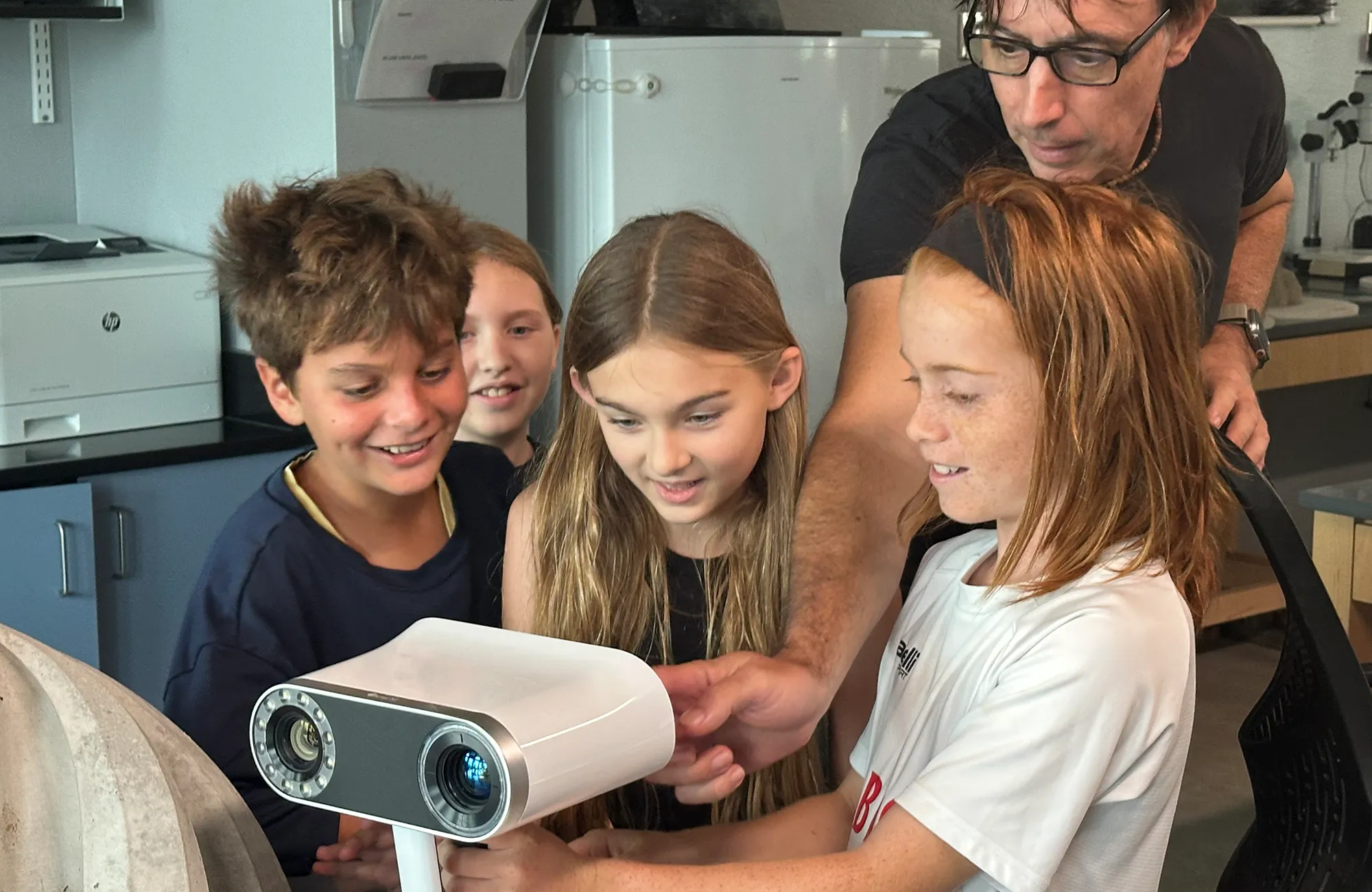 Four children and an adult examine a white 3D scanning device in a classroom or lab setting.